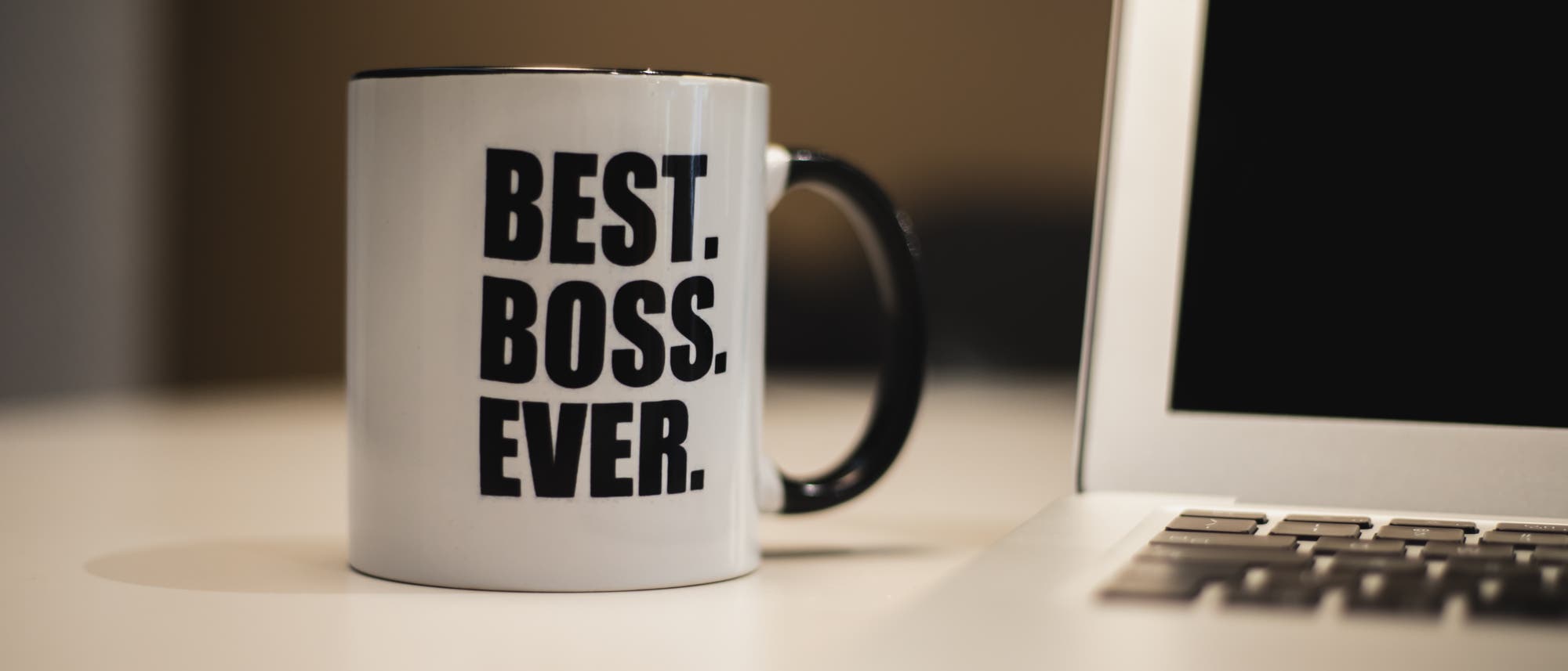 Eine Tasse mit der Aufschrift »Best. Boss. Ever.« steht auf einem Tisch neben einem Laptop Eine Tasse mit der Aufschrift »Best. Boss. Ever.« steht auf einem Tisch neben einem Laptop