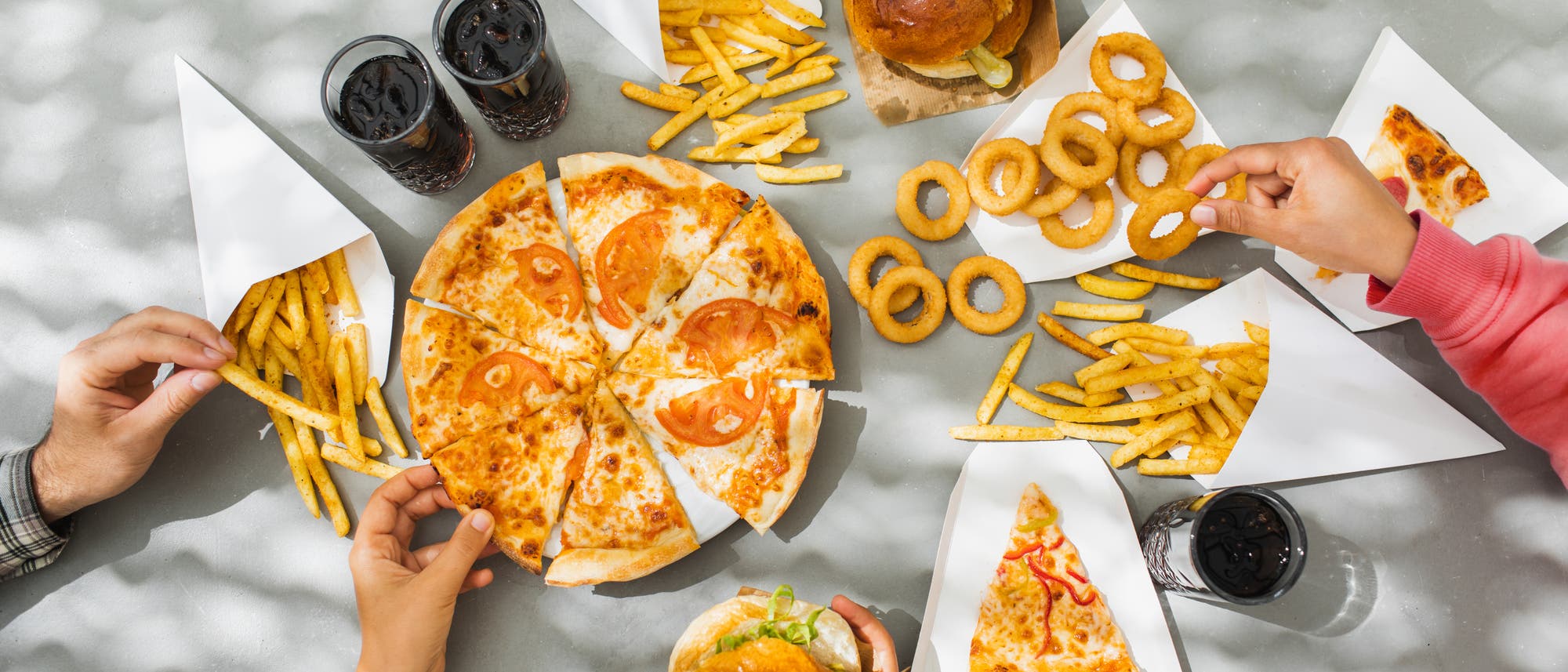 Eine Gruppe von Menschen teilt eine Mahlzeit auf einem Tisch. In der Mitte befindet sich eine Pizza mit Tomatenscheiben, umgeben von Pommes frites in Papiertüten, Zwiebelringen und einigen Gläsern Cola. Eine Person hält einen Zwiebelring, während eine andere ein Stück Pizza nimmt. Die Szene ist von Sonnenlicht durch Schatten von Blättern beleuchtet.