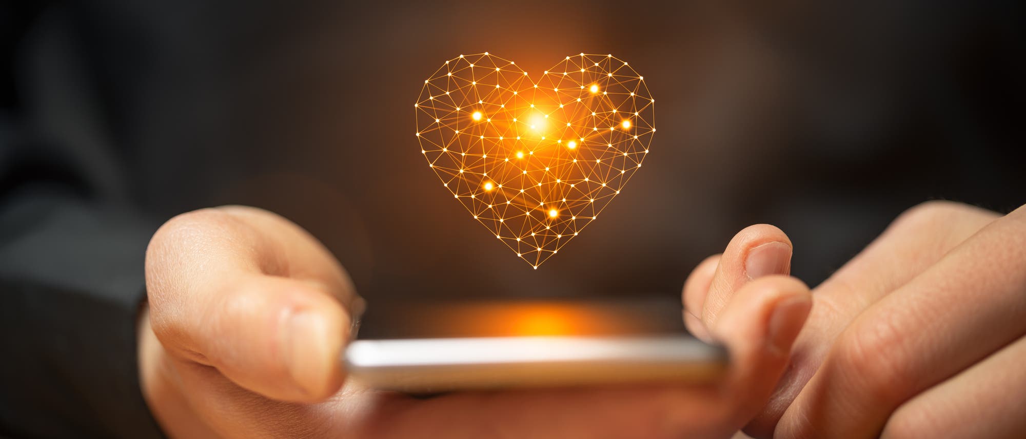 Eine Person hält ein Smartphone in der Hand, über dem ein leuchtendes, digitales Herz aus verbundenen Linien und Punkten schwebt. Das Bild symbolisiert digitale Kommunikation und Technologie. Der Hintergrund ist unscharf, um den Fokus auf das Herz und das Smartphone zu lenken.