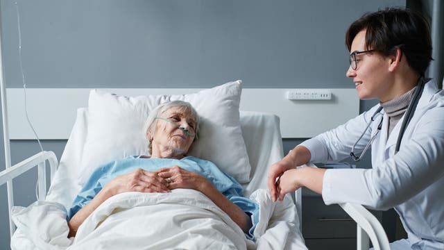 Eine ältere Frau liegt in einem Krankenhausbett, mit einer Sauerstoffbrille im Gesicht. Sie trägt ein hellblaues Krankenhaushemd und ist mit einer weißen Decke zugedeckt. Neben ihr sitzt eine Ärztin in einem weißen Kittel mit Stethoskop um den Hals, die freundlich mit der Patientin spricht. Die Szene vermittelt eine fürsorgliche medizinische Betreuung in einem Krankenhauszimmer.